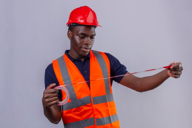 Young african american builder man wearing construction vest and safety helmet looking at using measuring tape looking at it standing 