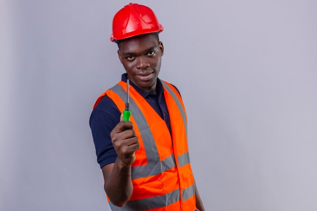 Young african american builder man wearing construction vest and safety helmet showing screwdriver with confident smile standing 