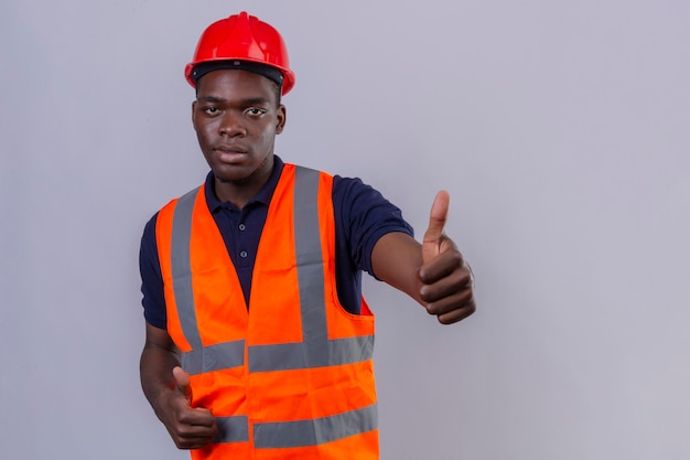 建設ベストと立っている親指を示す安全ヘルメットを身に着けている若いアフリカ系アメリカ人ビルダー男