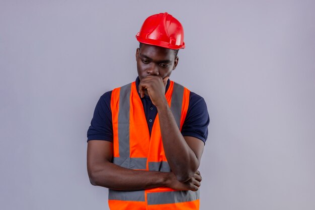 Young african american builder man wearing construction vest and safety helmet standing with hand on chin thinking trying to make choice worried standing 