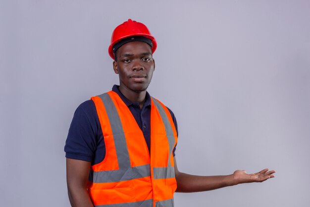 Молодой афро-американский строитель мужчина в строительном жилете и защитном шлеме с серьезным лицом, представляя руку и ладонь на изолированном белом