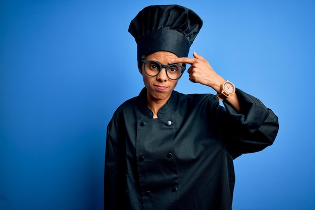 Foto gratuita giovane donna chef afroamericana che indossa l'uniforme da cucina e cappello su sfondo blu che punta infelice al brufolo sulla fronte brutta infezione di comedone acne e problema della pelle