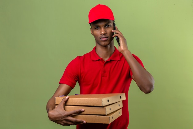 Young african american delivery man wearing red polo shirt and cap carrying stack of pizza boxes while speaking by smartphone over isolated green