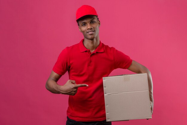 Молодой афроамериканец доставляющий носить красную рубашку поло и кепку, держа картонную коробку и указывая на коробку с пальцем, улыбаясь на изолированных розовый