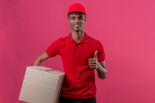 赤いポロシャツと段ボール箱を押しながら分離されたピンクの上に親指を現してキャップを身に着けている若いアフリカ系アメリカ人の配達人