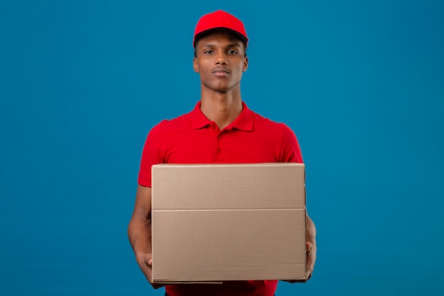 Free photo young african american delivery man wearing red polo shirt and cap holding cardboard box standing with serious face over isolated blue