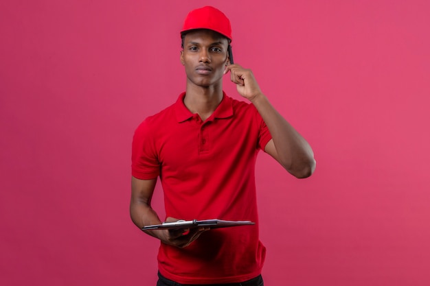 Молодой афроамериканец доставки человек, одетый в красную рубашку поло и шапку, держа буфера обмена и пятнышек на смартфон с серьезным лицом на изолированных розовый