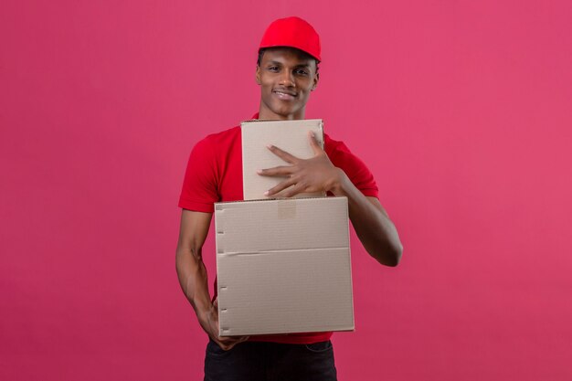 Young african american delivery man wearing red polo shirt and cap holding stack of boxes looking at camera with smile on face over isolated pink