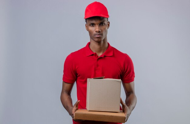 Молодой афроамериканец доставляющий носить красную рубашку поло и шапку, держа стопку картонных коробок, глядя на камеру с серьезным лицом над изолированной белой