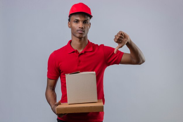 Young african american delivery man wearing red polo shirt and cap holding stack of cardboard boxes showing thumb down with serious face over isolated white