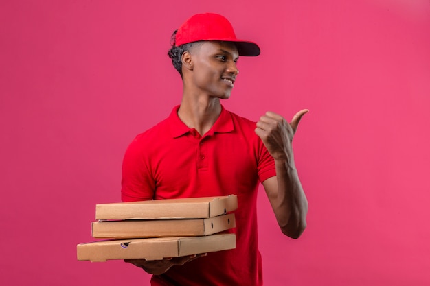 Молодой афроамериканец доставляющий носить красную рубашку поло и шапку, держа стопку коробок для пиццы, глядя в сторону, указывая пальцем в сторону с улыбкой на лице над изолированный розовый