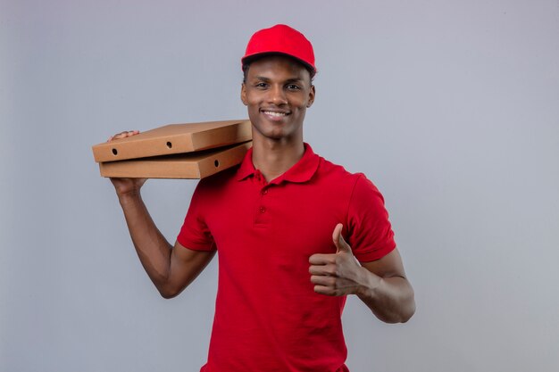 Молодой афроамериканец доставляющий носить красную рубашку поло и шапку, удерживая стопку коробок для пиццы, показывая большой палец вверх и улыбается над изолированный белый