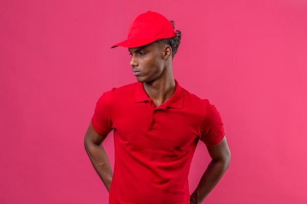 Free photo young african american delivery man wearing red polo shirt and cap looking away to side with serious face standing over isolated pink