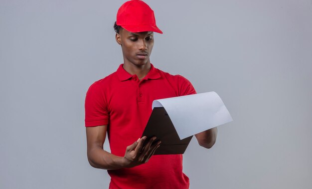 Young african american delivery man wearing red polo shirt and cap looking at clipboard with serious face over isolated white