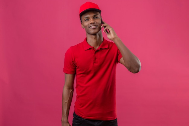 Young african american delivery man wearing red polo shirt and cap looking confident and speaking by smartphone over isolated pink