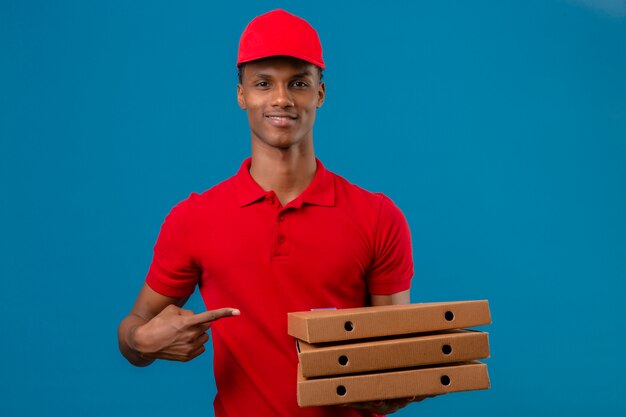 Молодой афроамериканец доставляющий носить красную рубашку поло и кепку, указывая пальцем на стопку коробок для пиццы в другой руке на изолированных синий