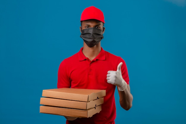 Young african american delivery man wearing red polo shirt and cap in protective mask and gloves standing with stack of pizza boxes showing thumbs up over isolated blue