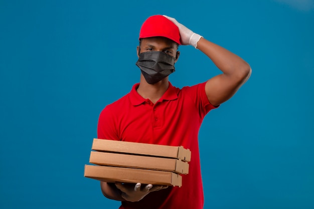 Young african american delivery man wearing red polo shirt and cap in protective mask and gloves standing with stack of pizza boxes surprised with hand on head for mistake bad memory concept