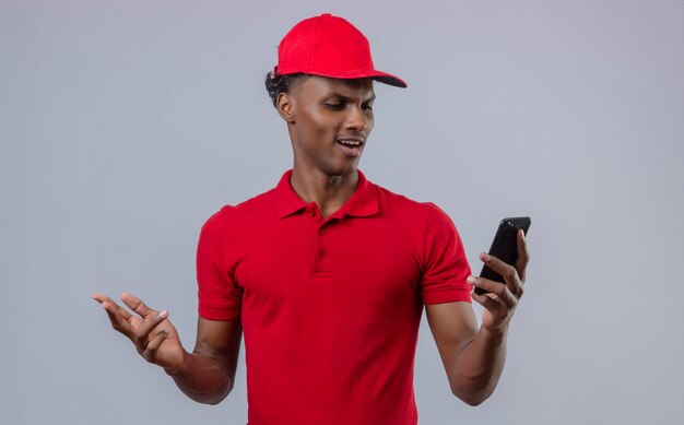 Молодой афроамериканец недоволен доставщиком в красной рубашке поло и кепке, глядя на смартфон в руке, показывая неприязнь над изолированной белой