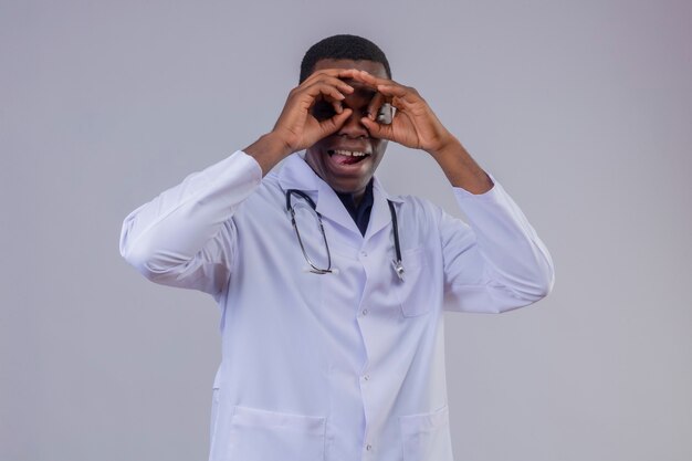 Молодой афро-американский врач в белом халате со стетоскопом делает нормальный жест, как бинокль, смотрит сквозь пальцы, высунув язык