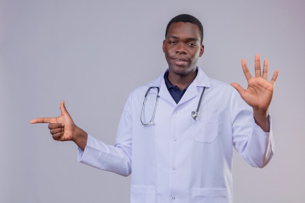 Young african american doctor wearing white coat with stethoscope looking confident showing number five and pointing with index finger to the side