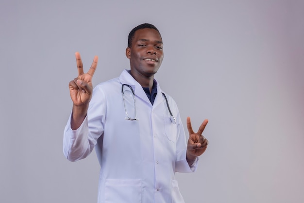 Young african american doctor wearing white coat with stethoscope showing victory sings with fingers of both hands smiling