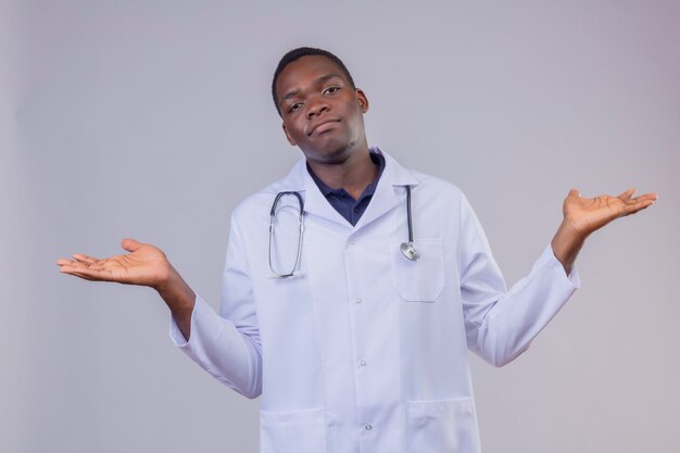 Young african american doctor wearing white coat with stethoscope shrugging shoulders, looking uncertain and confused, spreading palms