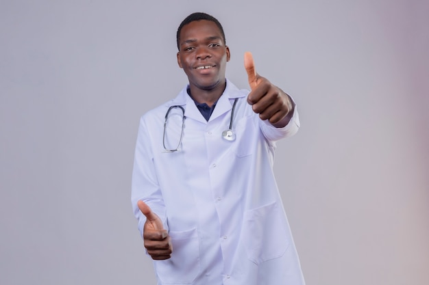 Молодой афро-американский врач в белом халате со стетоскопом, уверенно улыбаясь, показывает палец вверх обеими руками