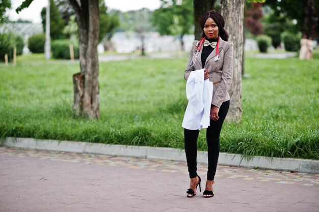 若いアフリカ系アメリカ人の女性医師は、屋外でポーズをとった聴診器で手に白いコートを保持します