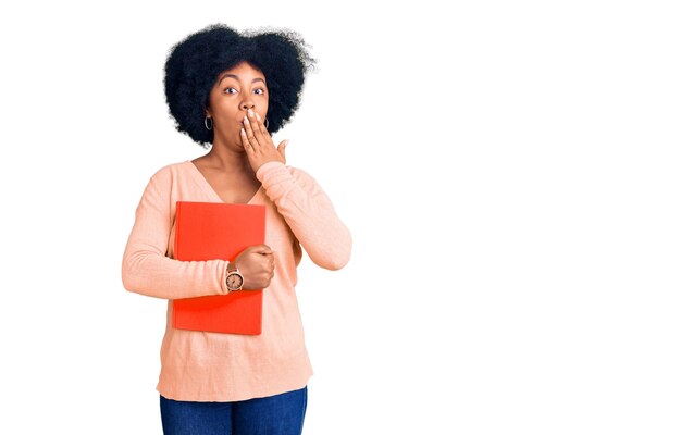 Young african american girl holding book covering mouth with hand, shocked and afraid for mistake. surprised expression