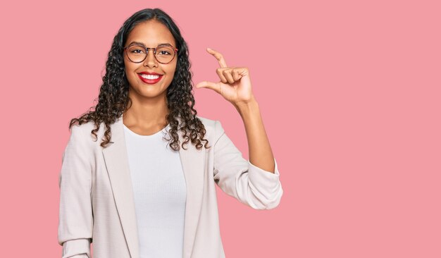 Young african american girl wearing business clothes smiling and confident gesturing with hand doing small size sign with fingers looking and the camera. measure concept.