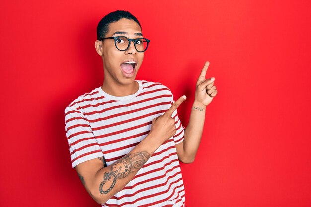 Young african american guy wearing casual clothes and glasses smiling and looking at the camera pointing with two hands and fingers to the side