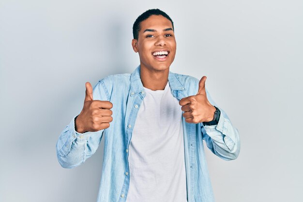 カジュアルな服を着た若いアフリカ系アメリカ人の男が笑顔で親指を立て、幸せな陽気な表情と勝者のジェスチャーをして肯定的なジェスチャーをする成功のサインをしている