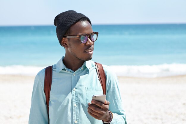 Young African American hipster typing text messages on smartphone while relaxing at seaside at daytime. Stylish black male using electronic gadget on beach, blue ocean and white sand in horizon