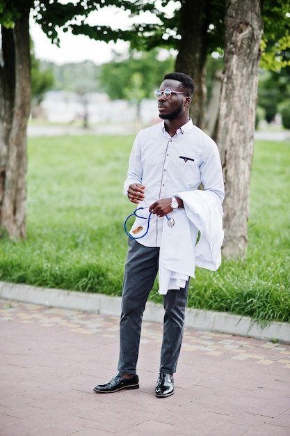 Молодой афроамериканец-врач держит под рукой белое пальто со стетоскопом на открытом воздухе