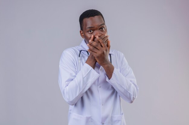 Young african american male doctor wearing white coat with stethoscope looking amazed and shocked covering mouth with hands