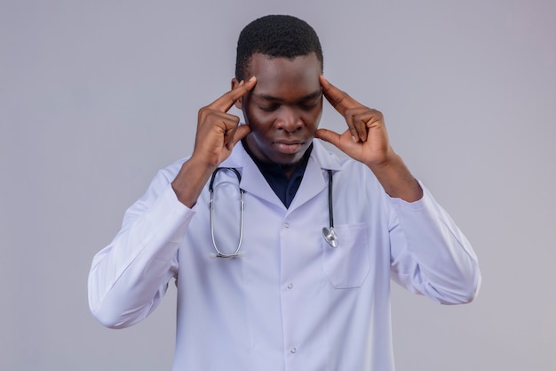 Giovane medico maschio afroamericano che indossa camice bianco con lo stetoscopio che sembra stanco di essere infastidito toccando le sue tempie con gli occhi chiusi