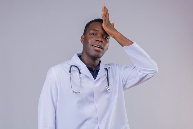 Giovane medico maschio afroamericano che indossa camice bianco con lo stetoscopio con la mano sulla sua testa per errore, dimenticato, concetto di cattiva memoria