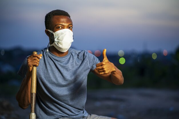 Foto gratuita giovane maschio afroamericano con una maschera protettiva che tiene in mano una pala e fa un gesto con il pollice in su