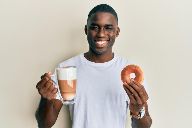 젊은 아프리카계 미국인 남자가 도넛을 먹고 얼굴에 행복하고 시원한 미소를 지으며 커피를 마시고 있습니다. 치아를 보여주는.