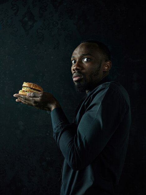 Молодой афроамериканец ест гамбургер и смотрит в сторону на черном фоне студии