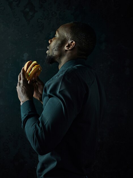 The young african american man eating hamburger and looking away on black studio