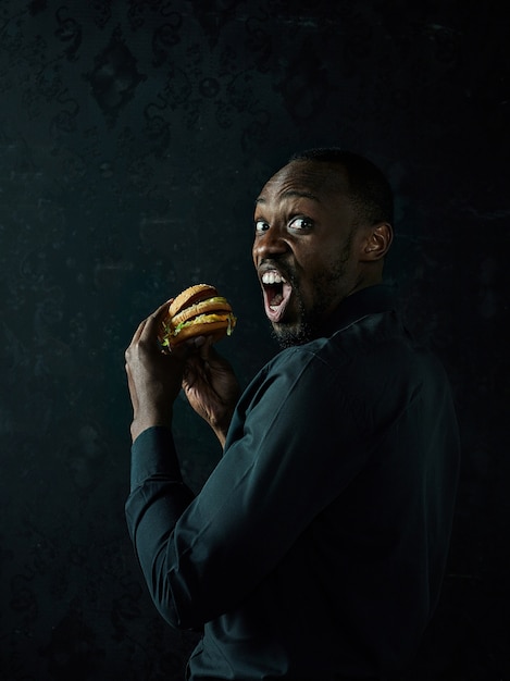 The young african american man eating hamburger and looking away on black studio