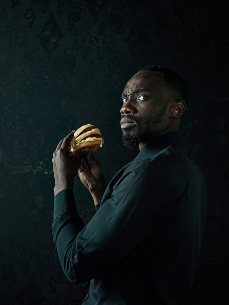 Free photo the young african american man eating hamburger and looking away on black studio