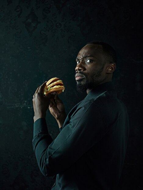 The young african american man eating hamburger and looking away on black studio