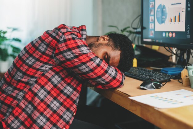 若いアフリカ系アメリカ人の男性フリーランサーが一生懸命働いて、自宅で仕事をしながら机の上で寝ているメンタルヘルスヘルスケアの概念