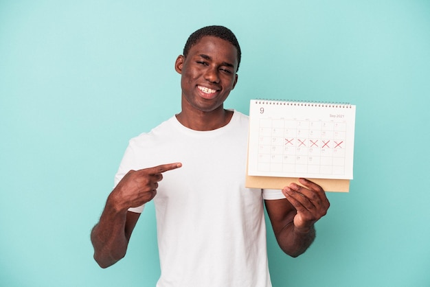 Young african american man holding a calendar isolated on blue background smiling and pointing aside, showing something at blank space.