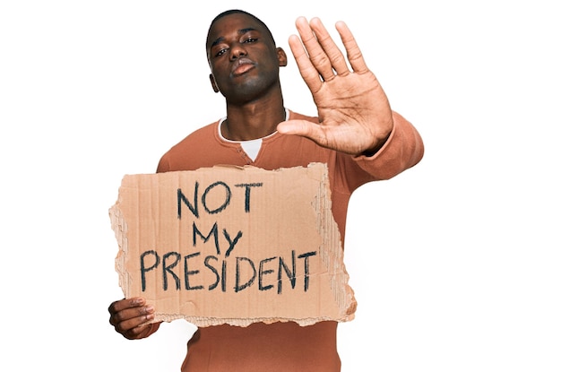 Молодой афроамериканец держит баннер протеста не моего президента с открытой рукой, делая знак "стоп" с серьезным и уверенным выражением лица в защиту жеста