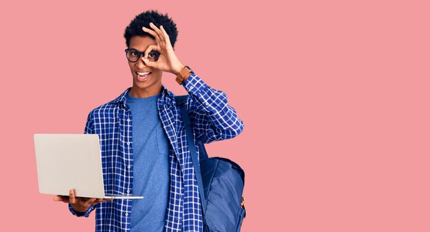 Молодой афроамериканец держит студенческий рюкзак с помощью ноутбука, улыбаясь, счастливо делая знак ок, положив руку на глаз, глядя сквозь пальцы