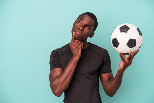 疑わしい​と​懐疑的​な​表現​で​横向き​に​見える​青い​背景​に​孤立した​サッカー​を​している​若い​アフリカ系​アメリカ人​の​男​。